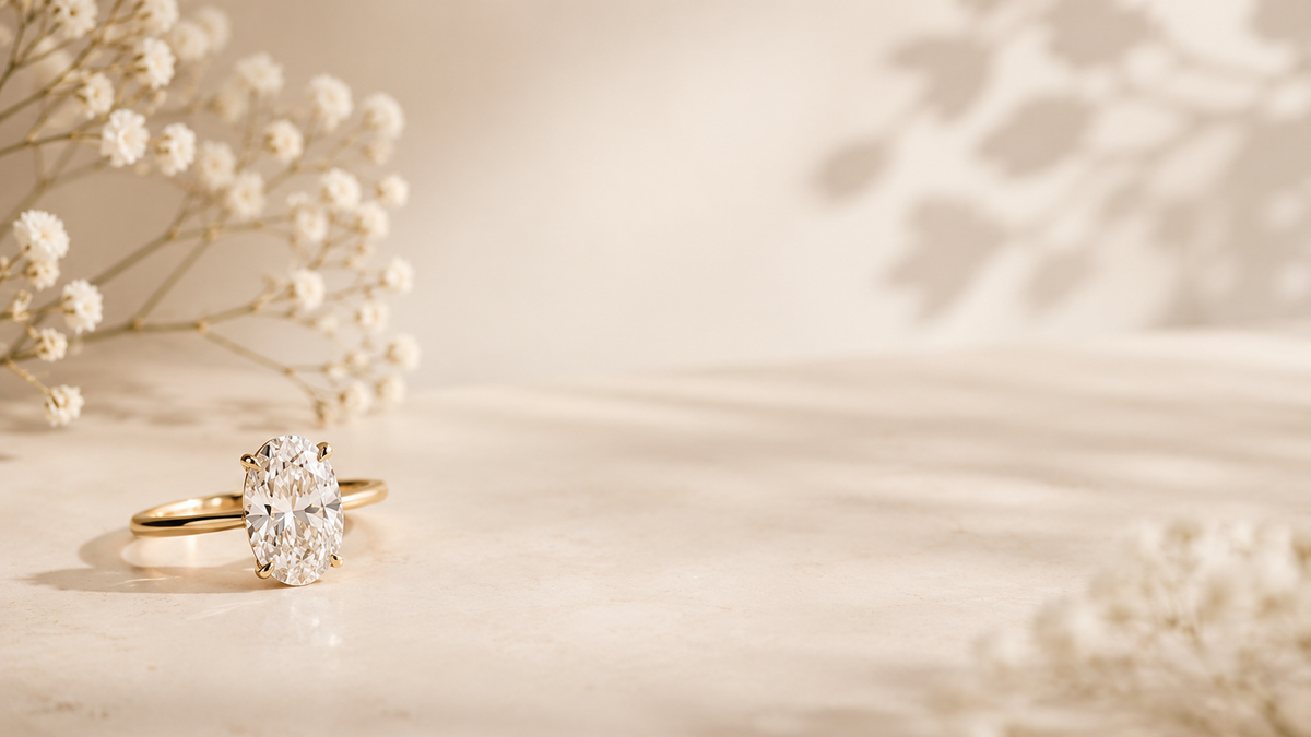 An Isla Bella gold oval solitaire engagement ring beside sprigs of baby's breath on soft champagne.